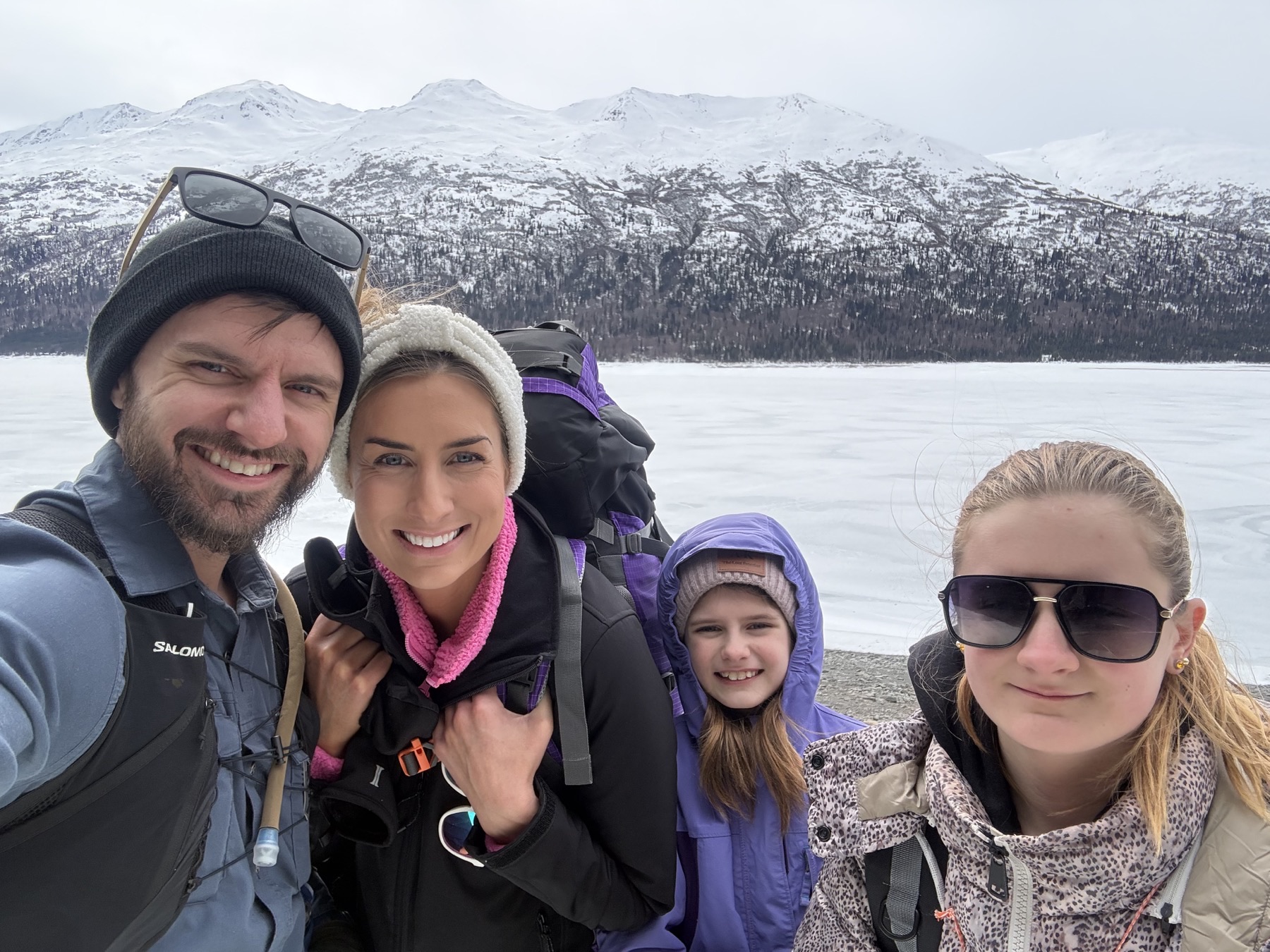 A family of four smiling together beside a frozen lake and snowy mountains.